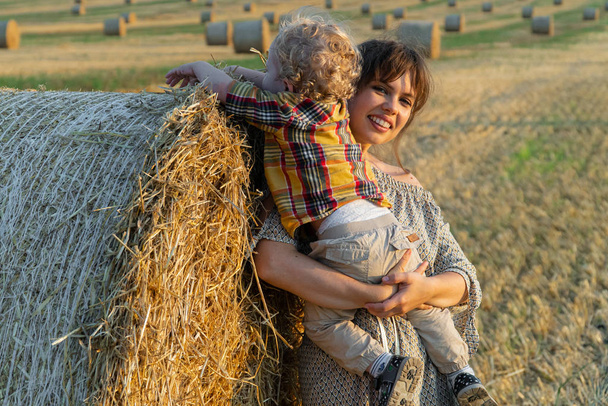 Young mother playing with her little son near a straw bale on a wheat field - Photo, Image