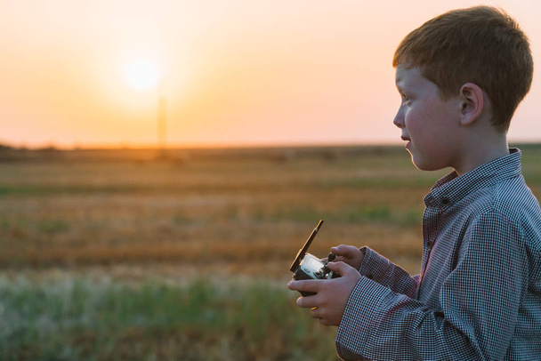 Red-haired boy at sunset in the evening stands on a wheat field with bales and controls the drone - Foto, afbeelding