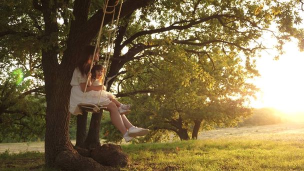 Maman secoue sa fille en se balançant sous un arbre au soleil. mère et bébé montent sur une balançoire à corde sur une branche de chêne dans la forêt. La fille rit, se réjouit. Amusement en famille dans le parc, dans la nature. chaude journée d'été
. - Photo, image