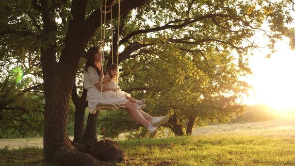 Moeder schudt haar dochter op schommel onder een boom in de zon. moeder en baby rijden op een touwschommel op een eiken tak in bos. Meisje lacht, verheugt. Familie plezier in Park, in de natuur. warme zomerdag. - Foto, afbeelding
