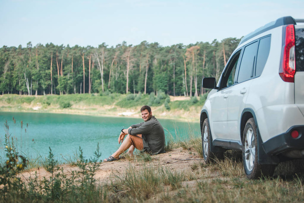 hombre sentado cerca de coche suv blanco en el borde mirando al lago con agua azul
 - Foto, imagen