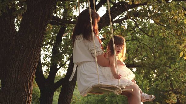 Mom shakes her daughter on swing under a tree in sun. close-up. mother and baby ride on a rope swing on an oak branch in forest. Girl laughs, rejoices. Family fun in park, in nature. warm summer day. - Photo, Image