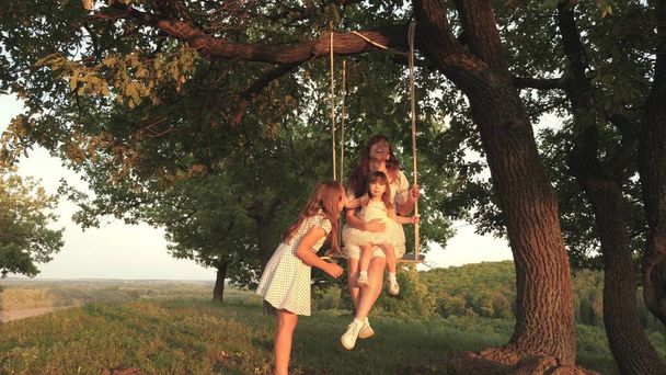 Mamá sacude a sus hijas en un columpio bajo el árbol bajo el sol, jugando con los niños. madre y el bebé montar en columpio cuerda en una rama de roble en el bosque. chica ríe, se regocija. Diversión familiar en el parque, en la naturaleza
 - Foto, imagen