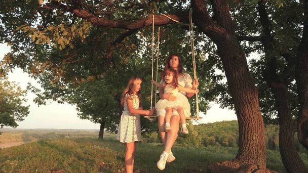 Η μαμά κουνάει τις κόρες της σε μια κούνια κάτω από το δέντρο στον ήλιο, παίζοντας με τα παιδιά. μητέρα και μωρό βόλτα με σχοινί swing σε ένα κλαδί βελανιδιάς στο δάσος. κορίτσι γελάει, χαίρεται. Οικογενειακή διασκέδαση στο πάρκο, στη φύση - Φωτογραφία, εικόνα