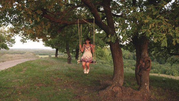 Młoda dziewczyna swinging na liny huśtawka na gałęzi dębu. Piękna dziewczyna w białej sukni w parku. Teen Girl cieszy lot na huśtawce na letni wieczór w lesie - Zdjęcie, obraz