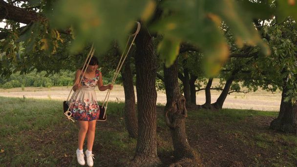 mooie tiener meisje geniet van een vlucht op een schommel op een zomeravond in het bos. gelukkig meisje swingend op een touwschommel op een eiken tak. jong meisje in een witte jurk in het Park. - Foto, afbeelding
