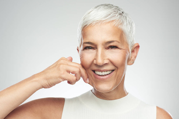 Gorgeous smiling Caucasian senior woman with short gray hair pinching her cheek. Beauty photography. - 写真・画像