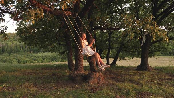 Młoda dziewczyna swinging na huśtawce pod drzewem w słońcu, bawiąc się z dziećmi. Zbliżenie. Rodzinna zabawa w przyrodzie. dziecko jeździ liny huśtawka na gałęzi dębu w parku zachód słońca. śmiech dziewczyna, raduje się. - Zdjęcie, obraz