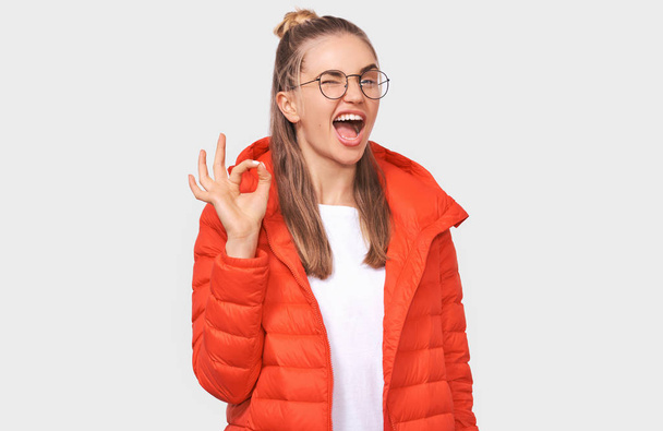 Retrato de una joven alegre con chaqueta roja, gafas transparentes, mostrando gestos Ok con la mano. Bastante hembra sonriendo ampliamente, posando sobre fondo blanco
. - Foto, imagen