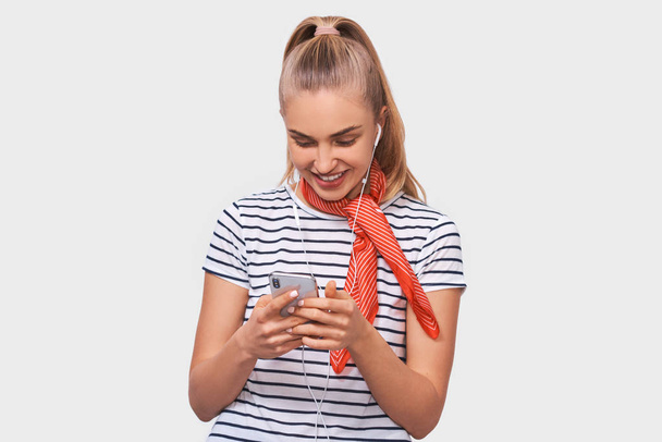 Joyful young woman with ponytail hairstyle, wearing striped t-shirt, stylish red scarf on neck, looking to the camera listening to her favourite songs via white earphones using smartphone - Fotografie, Obrázek