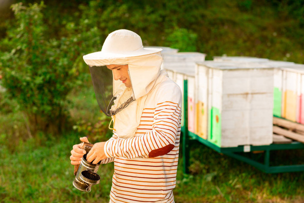 Un apicultor con una tapa protectora prepara una herramienta para fumar abejas de una colmena en el colmenar. Humo para abejas, equipo en colmenar
 - Foto, imagen