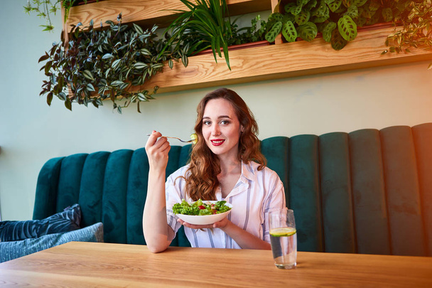 Joven mujer feliz comiendo ensalada en el hermoso interior con flores verdes en el fondo e ingredientes frescos en la mesa. Concepto de comida saludable
 - Foto, Imagen