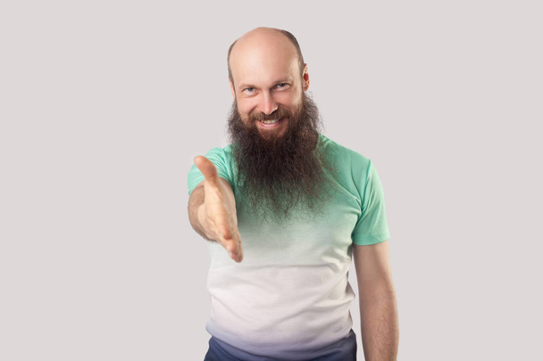 happy middle aged bald man with long beard in green t-shirt looking at camera with toothy smile and giving hand to handshake while greeting on grey background - Fotoğraf, Görsel