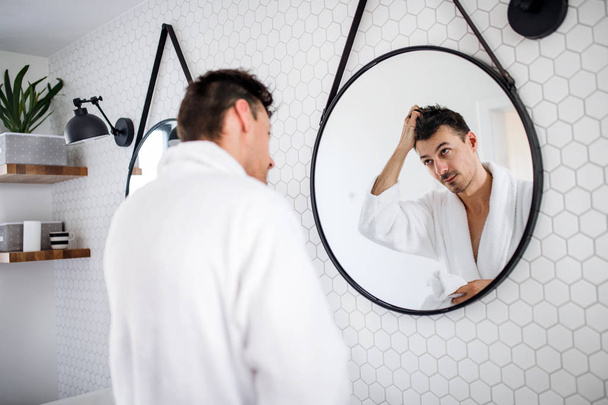 Young man looking in mirror in the bathroom in the morning, daily routine. - Фото, изображение