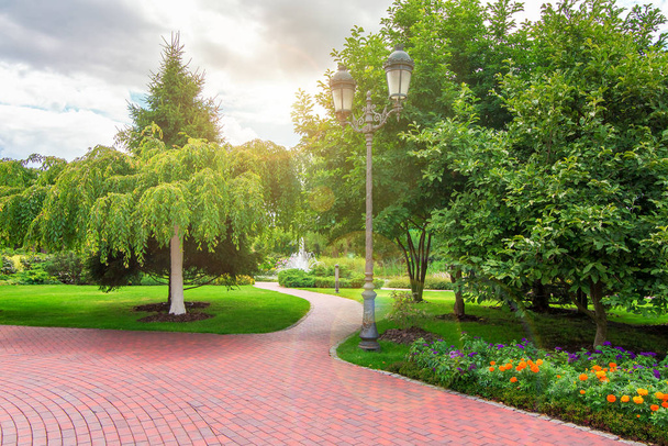 pedestrian sidewalk in a park with green plants and an iron retro lantern, trees and flowers in flower beds with sun flare. - Foto, afbeelding