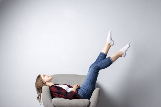 genç kız bacakları yukarı kaldırdı beyaz bir duvara karşı yumuşak rahat bir sandalyede oturuyor, bir hipster öğrenci dinlenme ve hayal, kopya alanı - Fotoğraf, Görsel