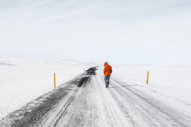 Wide shot of a male wearing orange jacket walking along a snowy highway road - Photo, Image