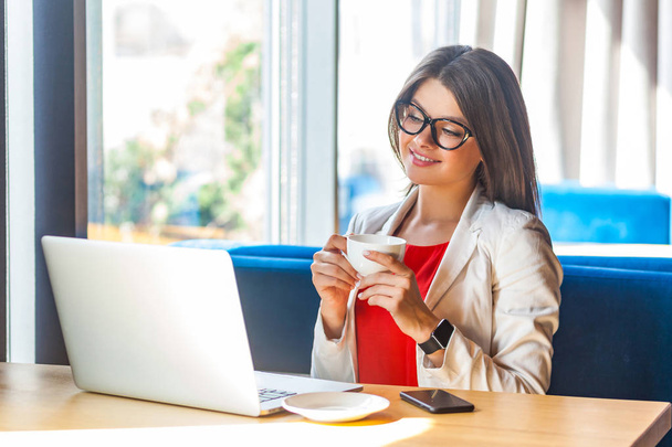 happy beautiful stylish brunette young woman in glasses holding cup of coffee and looking at laptop screen while sitting at table  in cafe, freelancing concept    - Valokuva, kuva
