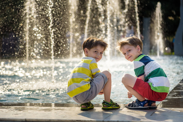 Cute toddler boy and older brothers, playing on a jet fountains with water splashing around, summertime - Photo, Image