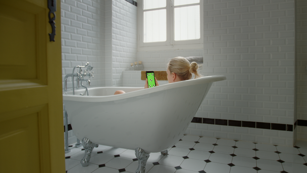 Gyönyörű szőke lány vesz egy Bath használ zöld modell-up Screen smartphone pihenés közben. Vonzó lány használ chroma kulcs mozgatható telefon részére szemöldök társadalmi média, néz video, szörfözés Internet - Felvétel, videó