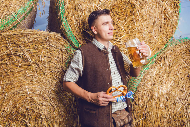 young man in traditional german costume holding beer bug with pretzel and standing near haystacks - Фото, зображення