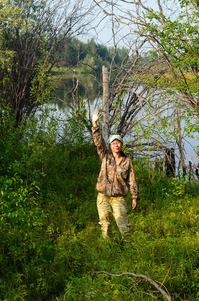 Yakut ragazza asiatica turista vestito con abiti protettivi di colore agitando la mano in piedi sulla riva del lago selvaggio invaso nella foresta
. - Foto, immagini