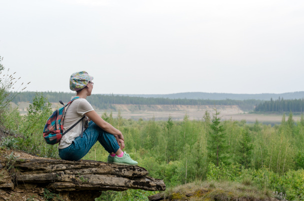 Yakut Asian girl tourist with a backpack and a cap sitting on the edge of the mountain in the wild tundra of Yakutia hugging his legs resting and looking into the distance to the river vilyu. - Photo, image