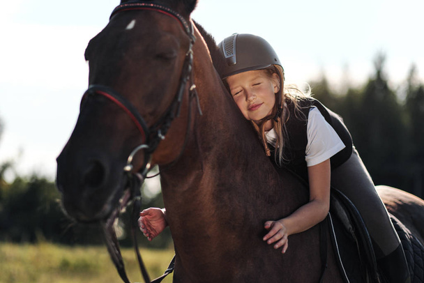 Girl teenager jockey sits on a brown horse, hugs and strokes her. - Fotoğraf, Görsel