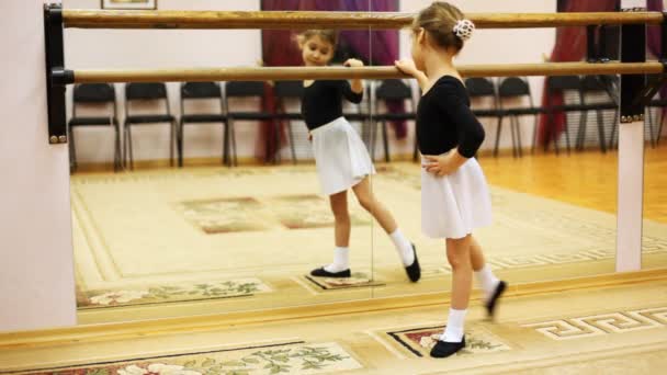 Little girl does ballet leg movements while standing sideways to mirror at hall - Filmmaterial, Video