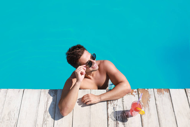 Handsome young man with refreshing cocktail in swimming pool on sunny day, above view - Foto, imagen