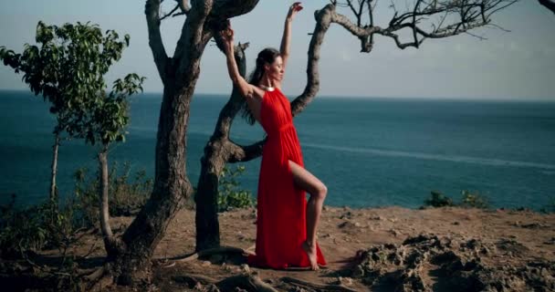 Gyönyörű nő piros ruhában pózol a tenger partján szikla alatt napsütéses nyári napon videó lassítva - Felvétel, videó