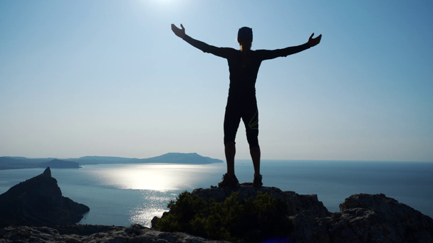 若い長い髪の男は、美しい青空に対して海の上の山の頂上に立って手を上げます。頂上に立つ幸せなハイカー男のシルエット. - 映像、動画