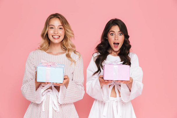 Happy young two pretty women girls friends sisters posing isolated over pink wall background holding present boxes. - Fotoğraf, Görsel