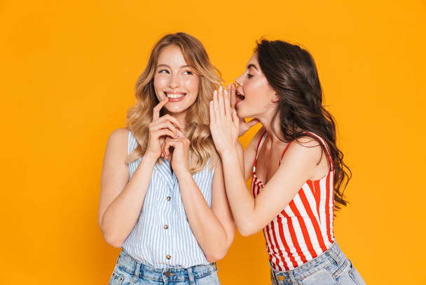Portrait of two seductive blonde and brunette women 20s in summer wear expressing delight while whispering gossips - Fotó, kép