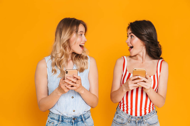 Portrait of two delighted blonde and brunette women 20s in summer wear smiling while holding cellphones - Foto, afbeelding