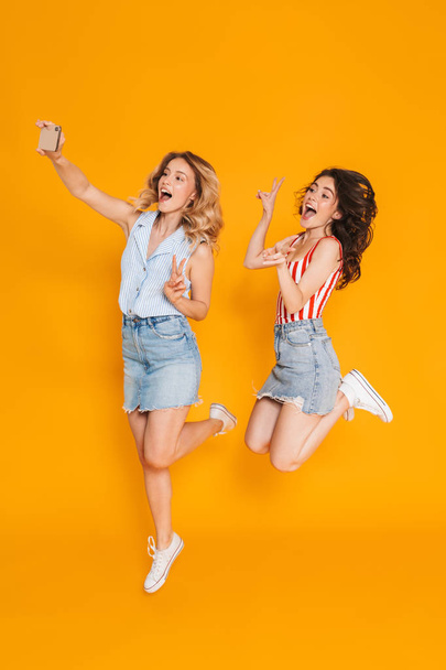 Portrait of two delighted blonde and brunette women gesturing peace sing and jumping while taking selfie photo on cellphone - Φωτογραφία, εικόνα