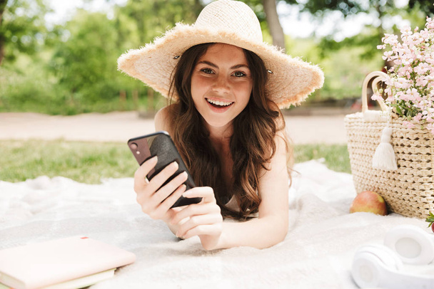 Photo of beautiful young woman wearing straw hat holding smartphone while having picnic in green park - Zdjęcie, obraz
