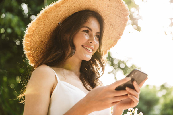 Photo of happy elegant caucasian woman wearing straw hat using cellphone while walking in green park - Zdjęcie, obraz