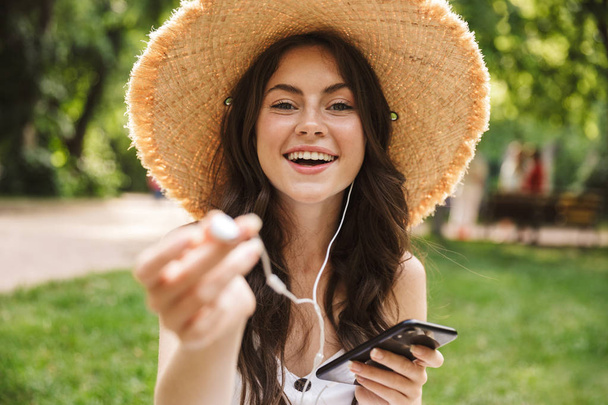 Excited happy young pretty woman outoors in green park in hat sitting on grass using mobile phone listening music. - 写真・画像