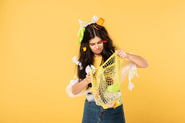 confused young woman with trash in hair holding bag with rubbish isolated on yellow - Foto, immagini
