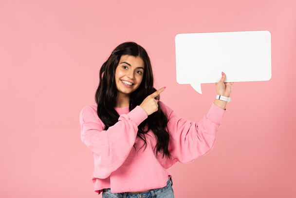 χαμογελαστή κοπέλα που δείχνει την κενή φούσκα ομιλίας, απομονωμένη σε ροζ - Φωτογραφία, εικόνα