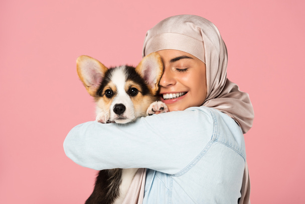 Veselá Arabská žena v hidžábu, která drží štěně Welsh Corgi, izolovaná na růžovém - Fotografie, Obrázek