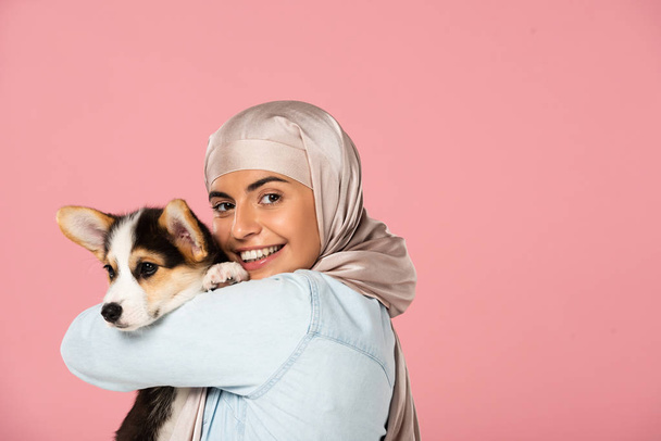 コルギーの子犬を持つヒジャーブで笑顔のイスラム教徒の女性, ピンクで隔離 - 写真・画像
