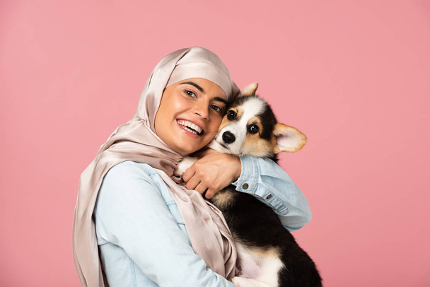 beautiful arabic girl in hijab holding Welsh Corgi puppy, isolated on pink - Photo, image