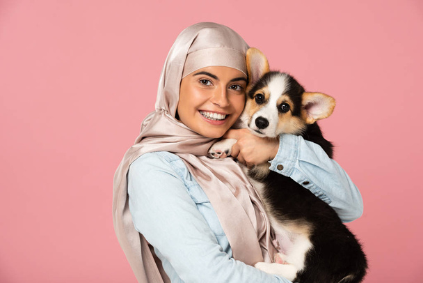 ピンクに隔離されたウェールズコーギーの子犬を抱いてヒジャーブで笑顔のアラビアの女の子 - 写真・画像