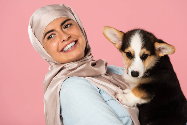 beautiful smiling muslim woman in hijab holding Welsh Corgi puppy, isolated on pink - Photo, Image