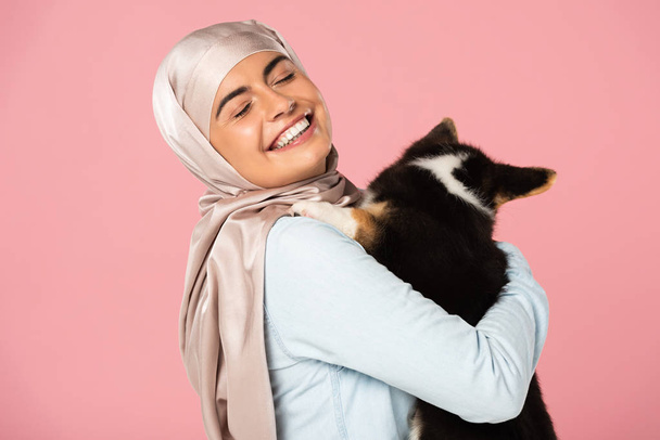 cheerful arabian girl in hijab holding cute puppy, isolated on pink - Zdjęcie, obraz