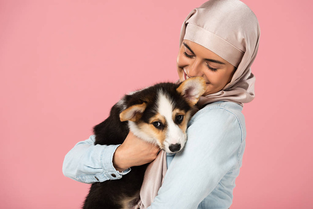 happy muslim girl in hijab holding cute corgi puppy, isolated on pink - Foto, Imagem