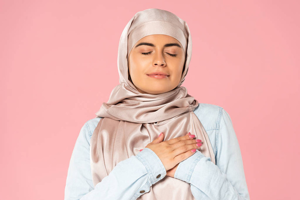 dreamy muslim girl holding hands on heart, isolated on pink - Fotografie, Obrázek
