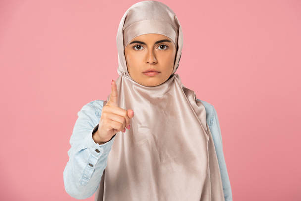 σοβαρή αραβική γυναίκα στη μαντίλα που σας σημαδεύει, απομονωμένη σε ροζ  - Φωτογραφία, εικόνα
